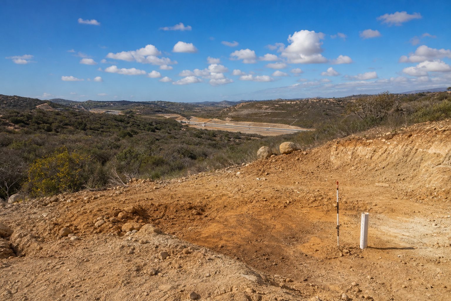 Grading study area on vacant Otay Ranch lot showing exposed soil layers and Otay Lakes Road infrastructure in the background.